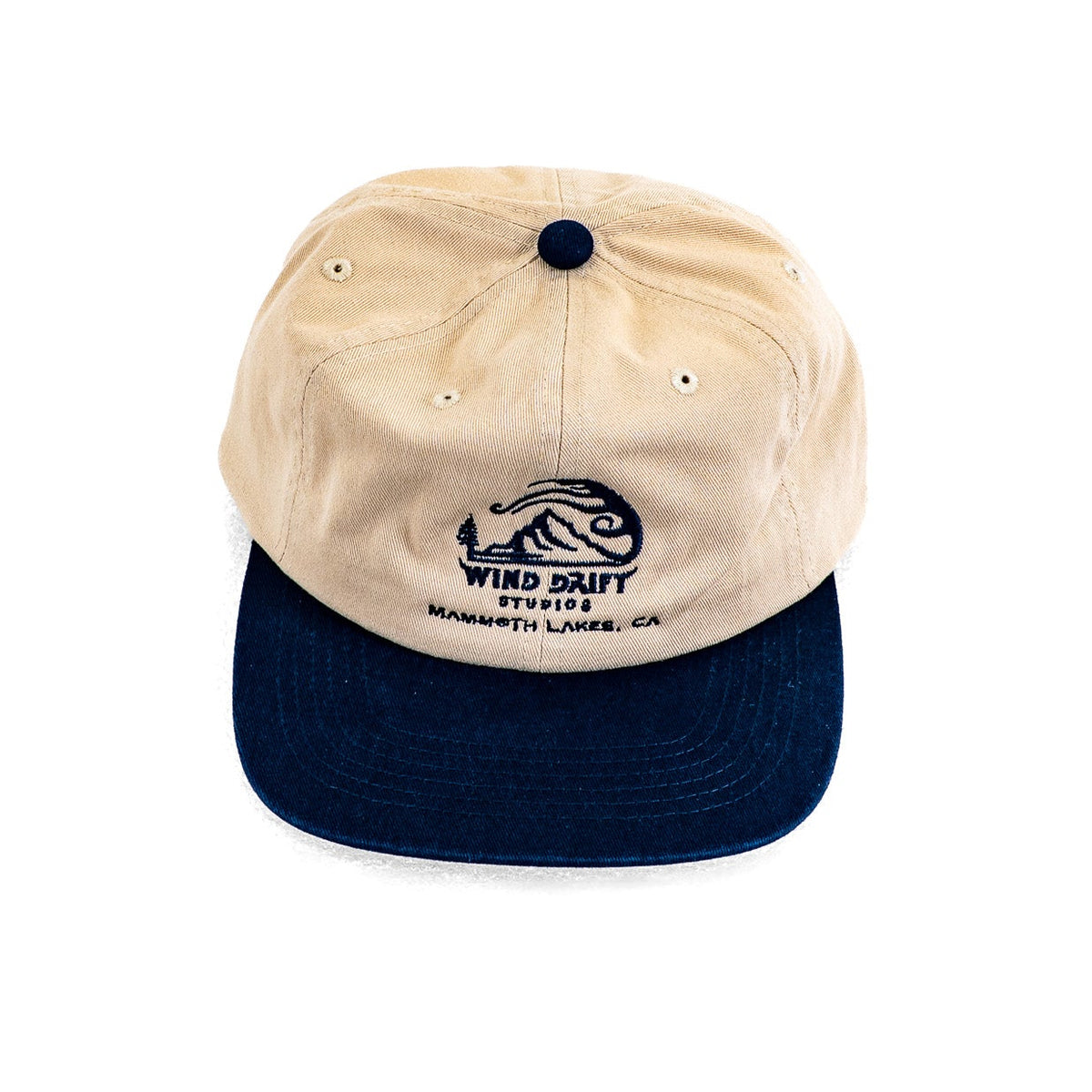 A two-tone baseball cap with a beige cotton front and a dark navy blue flat brim, featuring a navy blue embroidered Wind Dust Studios logo on the front.